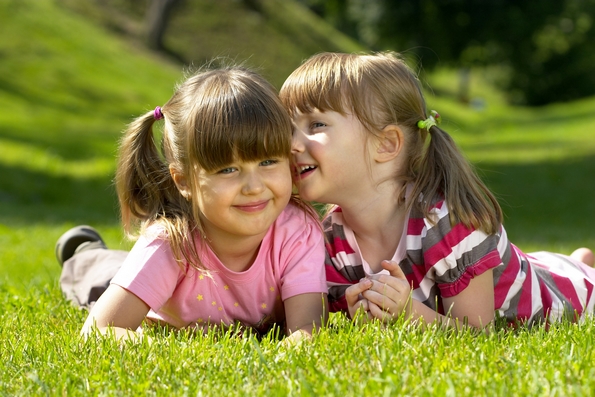 Two little girl lying on the grass in the park. One whispering a secret to another.